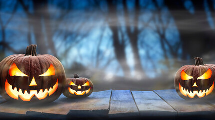 Three spooky halloween pumpkins, Jack O Lantern, with evil face and eyes on a wooden bench, table with a misty night forest background with space for product placement.