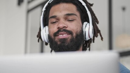 Wall Mural - A close-up view of a smiling young african american man wearing headphones is using his silver laptop computer in the living room at home