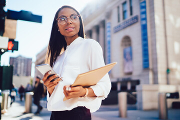 Charming black woman in glasses browsing smartphone with documents in hand and looking away in sunny street