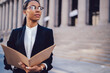 Thoughtful dark skinned female financial manager in black suit holding folder in hands while standing against office building outdoors. African American student of high financial economist university