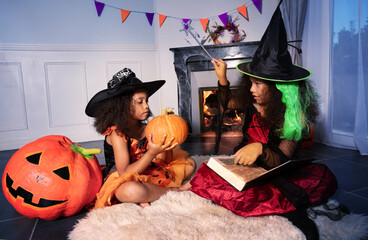 Two girls with trick or magic stick hold book in Halloween dress and cast a spell on orange pumpkin sitting near fireplace