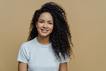 Horizontal Shot Of Curly Haired Woman With Healthy Skin, Toothy Smile, Enjoys Being Photographing, Wears Casual White T Shirt, Isolated On Beige Background. People, Emotions And Lifestyle Concept