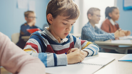 In Elementary School Class: Portrait of Brilliant Caucasian Boy Writes in Exercise Notebook, Taking Test and Writing Exam. Diverse Group of Bright Children Working Diligently and Learning