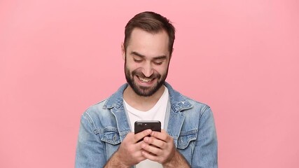 Wall Mural - Dreamful happy bearded young man guy 20s years old in denim jacket white t-shirt using mobile cell phone send sms girlfriend chatting isolated on pastel pink background studio People lifestyle concept