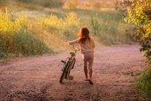 Little Girl Goes With A Bicycle On A Rural Road In Nature At Sunset