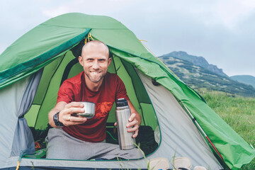 Wall Mural - smiling man in the green tent drinking the tea from a thermos with mountain background. he offering 