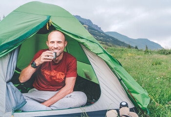 Wall Mural - sincerely smiling man in the green tent drinking the coffee from a thermos with mountain background 