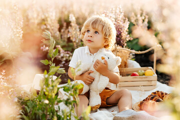 Little blonde boy wearing a straw hat, sitting in a field at sunset, feeding a plush bear toy a croissant. Cute kid at a picnic.