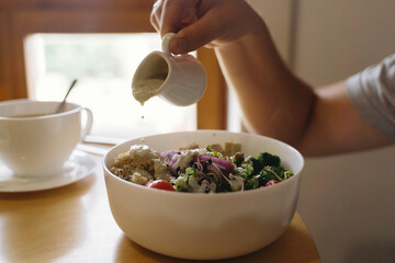 Buddha bowl, healthy eating concept. Man pouring sauce on vegan meal in restaurant. Bowl with avocado, radish, broccoli, tofu, tomato, couscous, microgreen and sesame seeds.