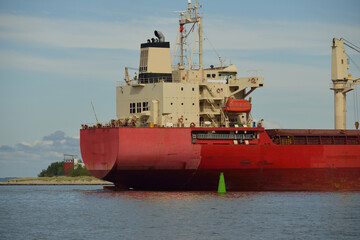 Wall Mural - Large red bulk carrier (cargo ship) with a cranes arriving to the Europoort. Green navigational buoy close-up. Rotterdam, Netherlands. Global communications, logistics, industry theme