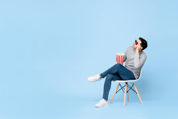 Young Caucasian man with popcorn and 3d movie glasses looking at empty space while sitting on chair isolated on light blue background