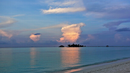 Magical sunset in the Maldives. The aquamarine ocean is calm. The colors of the sky are azure, blue, lilac. Clouds glow in the rays of the setting sun in gold and pink. Reflection in water. 