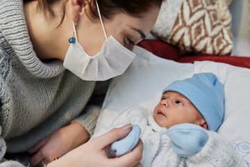 young redheaded mother with preventive health mask and lgtbiq+ bracelet, next to her newborn baby playing and laughing together on the bed