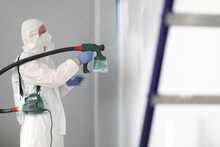 Close-up Of Professional Male Worker Painting Wall In Grey Colour With Spray Gun. Adult Man In White Protective Costume And Face Mask. Black Metal Ladder In Right Side. Renovation Concept