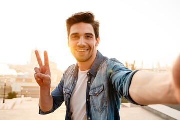 Poster - Photo of joyful young man gesturing peace sign while taking selfie