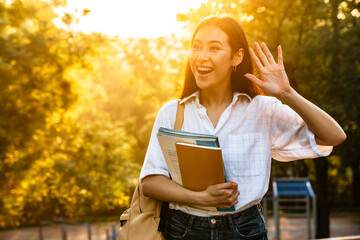 Wall Mural - Photo of happy asian student woman waving hand while walking in par