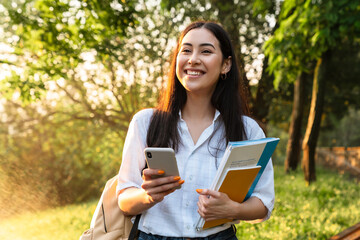 Wall Mural - Photo of happy asian student woman using cellphone while walking in parkPhoto of happy asian student woman with exercise books using cellphone while walking in green park