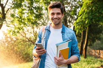 Wall Mural - Photo of smiling student man using smartphone while walking in park