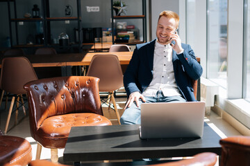 Caucasian young businessman wearing fashion suit is talking on mobile phone in modern office room at the desk in modern office on background of large window, laptop on table. Concept of office working