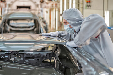 An employee of the car body paint shop with a medical mask on his face checks the quality of the painted surface