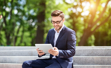 Business executive sitting on stairs in city park and browsing net on his tablet computer