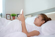 © PBXStudio - Asian woman is playing a smartphone on a bed in a room.