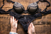 Woman Is Holding In Hands A Black Lingerie Panties Close Up On A Brown Store Counter Flat Lay Background.