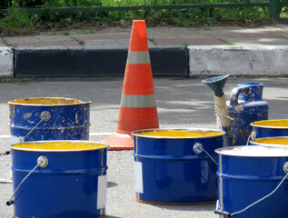 Repair work on the road. Blue large cans with white and yellow paint, next to it is an orange traffic signal cone