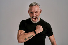 Middle Aged Muscular Man In Black T Shirt Shouting At Camera, Beating His Chest While Posing In Studio Over Grey Background