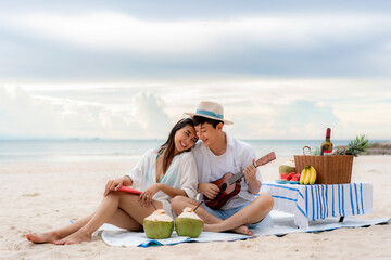 Asian couple happy and enjoy the trip honeymoon of couple lover on the sea beach by playing Ukulele and singing song music together. Summer, holidays, vacation and happy people in Thailand concept.