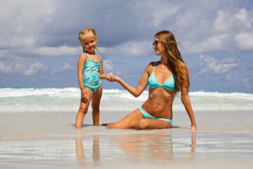 Wall Mural - Beautiful sexy young caucasian woman in blue bikini and little girl on the sandy beach. Summer family vacations, travel and tourism concept after end of coronavirus covid-19 lockdown. Copy space