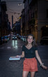 Young Asian teen in black blouse standing on stone street in front of building in black blouse at night holding box of pizza