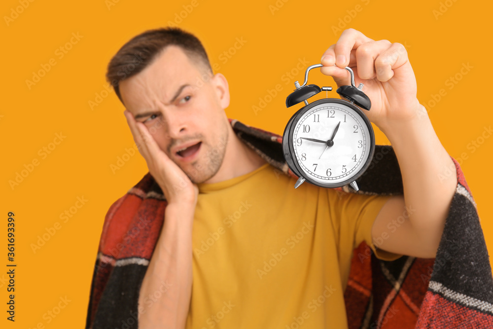Displeased man with alarm clock and plaid on color background. Concept of sleep deprivation