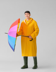 Wall Mural - Young Asian man in raincoat and with umbrella on grey background