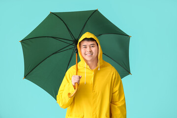 Wall Mural - Young Asian man in raincoat and with umbrella on color background