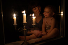 Mother And Daughter At The Window Look At The Burning Candles And Talk. The Girl Shows The Candles On Fire. Sparkle In The Eyes