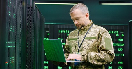 US army senior Caucasian male soldier working at laptop computer in dark room with monitors. Technician from military forces typing on keyboard and sending secret data of surveillance.