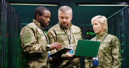 Male and female multiethnic army officers standing in monitoring analytic cener with servers with laptop computer. Woman and two men talking and discussing cybersecurity warfare strategy.