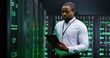 © VAKSMANV - African American man, data base administrator in server room, tapping on tablet device and checking information on computer. Work with secret information. Big data storage. Digital defence concept.