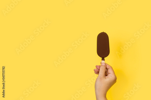 Fotomural Mano sosteniendo un helado de chocolate de palo batuta sobre un fondo amarillo brillante liso y aislado