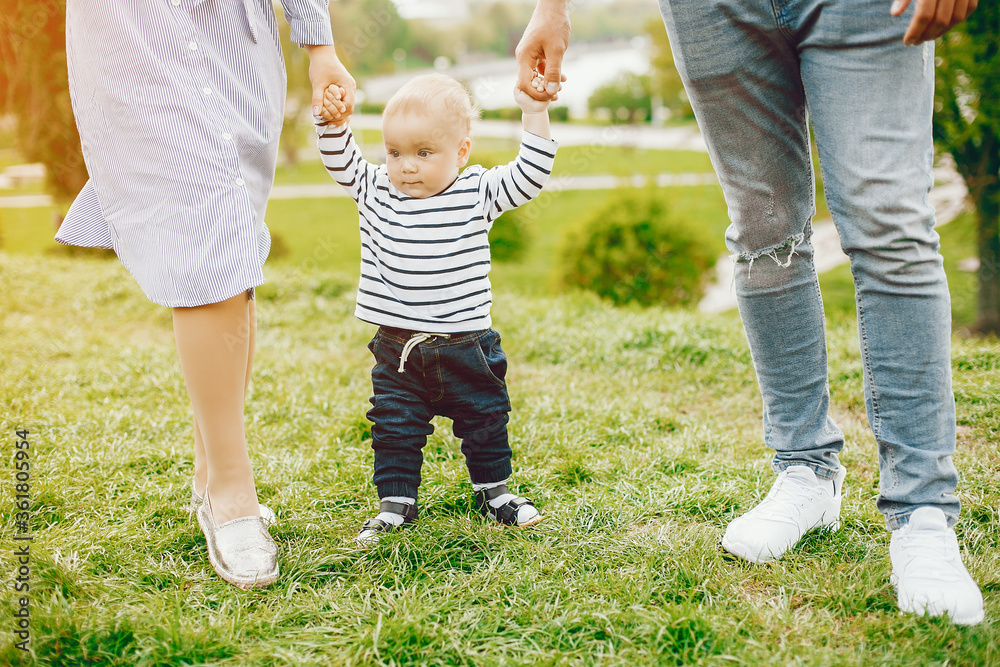 A young and beautiful blonde mother in a blue dress, along with her handsome man dressed in a white jacket, walks with her little son in the summer solar park - obrazy, fototapety, plakaty 