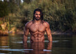 Leinwandbild Motiv Long haired bearded muscular man shirtless stands waist deep in the water. Aquaman in the water. Tanned attractive guy with long dark wet hair