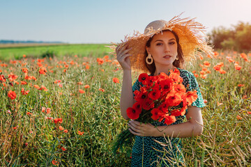 Wall Mural - Stylish woman holding bouquet of poppies flowers walking in summer field. Fashionable girl wearing straw hat