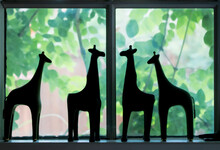 Cute Silhouettes Of Giraffe Figures Standing On A Window Ledge With Blurred Greenery And Flowers Showing Through