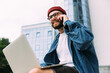 © mykolastock - Closeup portrait of modern bearded male hipster in eyeglasses talking on phone and using laptop at the same time