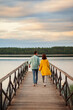 © Comeback Images - Rear perspective view of young romantic couple walking on wooden pier over river holding hands in summer evening