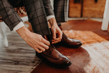 Groom In Black Grey Checkered Wedding Suit, White Shirt, Sitting On The Chair And Tied Shoelaces On Brown Leather Shoes. Business Clothing, Men's Style.