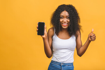 Portrait of a smiling young african american black woman holding blank screen mobile phone isolated over yellow background. Thumbs up.