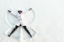 Black Girl In Fur Coat And High Top Boots Lies On White Glade And Draws Snow Angel Picture With Arms And Legs Top View