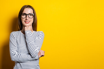 Friendly young woman with glasses holding a fist at his chin on a yellow background